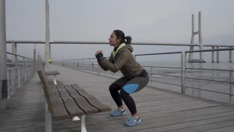 Slow-motion-shot-of-concentrated-young-woman-doing-squats-on-wooden-pier